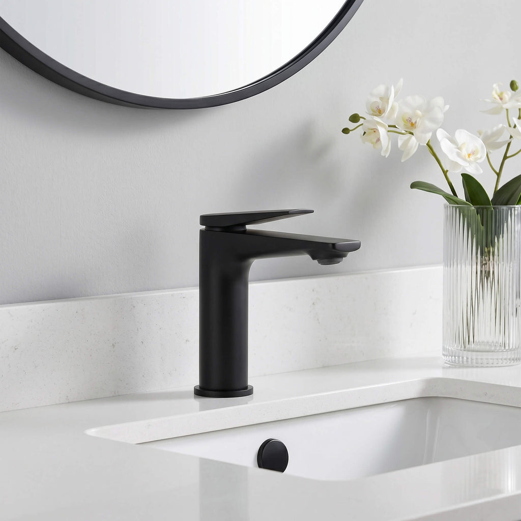 A matte black single-handle bathroom faucet installed on a white sink countertop, with a white wall, a round mirror, and a glass vase of white flowers in the background.