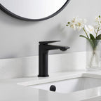 A matte black single-handle bathroom faucet installed on a white sink countertop, with a white wall, a round mirror, and a glass vase of white flowers in the background.